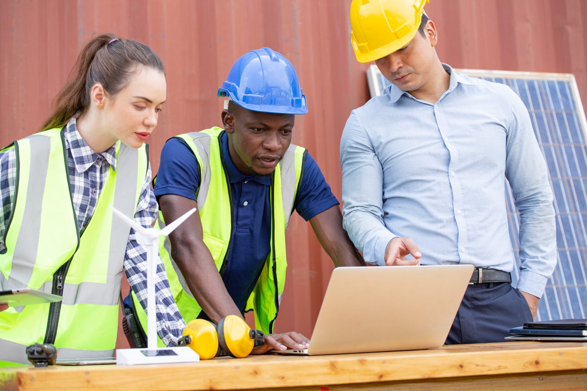 Three professionals in safety gear collaborating over a laptop at a construction site, highlighting hands-on apprenticeship training.
