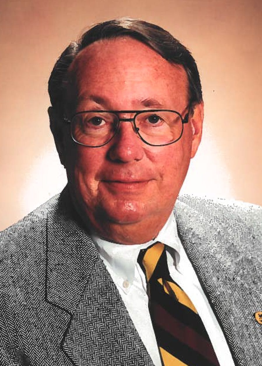 Professional headshot of an older man wearing glasses, a gray patterned jacket, white shirt, and striped tie, smiling against a beige background.