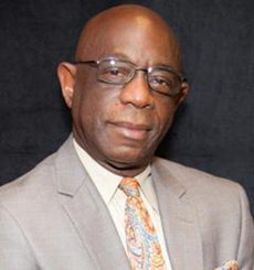 Professional headshot of a man wearing glasses and a light gray suit with a patterned tie, smiling slightly against a dark background.