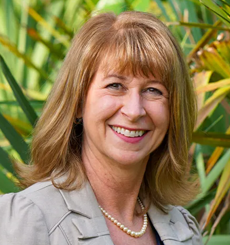 Professional headshot of a woman with light brown hair wearing a beige blazer and pearl necklace, smiling outdoors with green plants in the background.