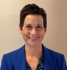 Professional headshot of a woman with short dark hair wearing a blue blazer and floral blouse, smiling against a beige wall.