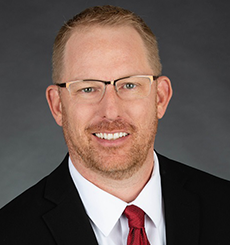 Professional headshot of a man with light brown hair and glasses wearing a dark suit and red tie, smiling against a dark gray background.