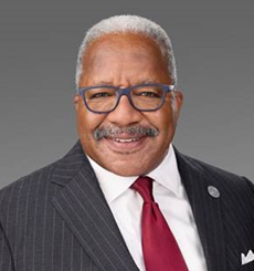 Professional headshot of a man with gray hair and mustache wearing a pinstripe suit, red tie, and glasses, smiling against a dark gray background.