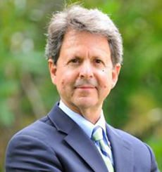 Professional headshot of a man with gray hair wearing a navy suit and green striped tie, standing outdoors with greenery in the background.