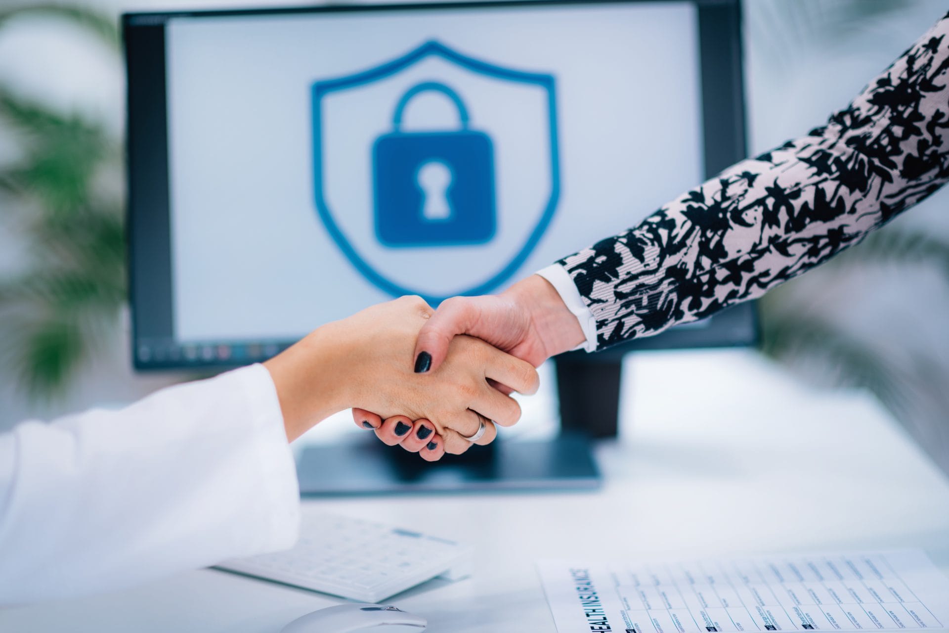 Two people shaking hands at a desk, with a computer screen in the background displaying a security lock icon, symbolizing privacy and data protection.