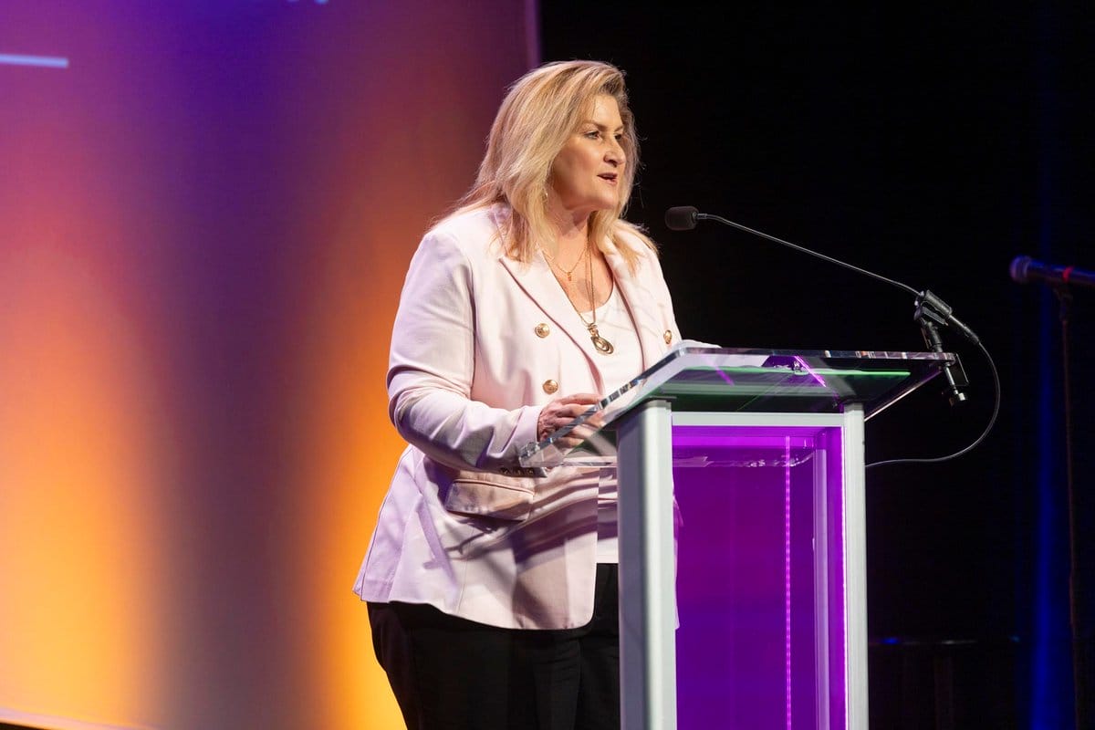 Professional woman speaking at a podium on stage with colorful lighting, symbolizing leadership and purpose in workforce development.