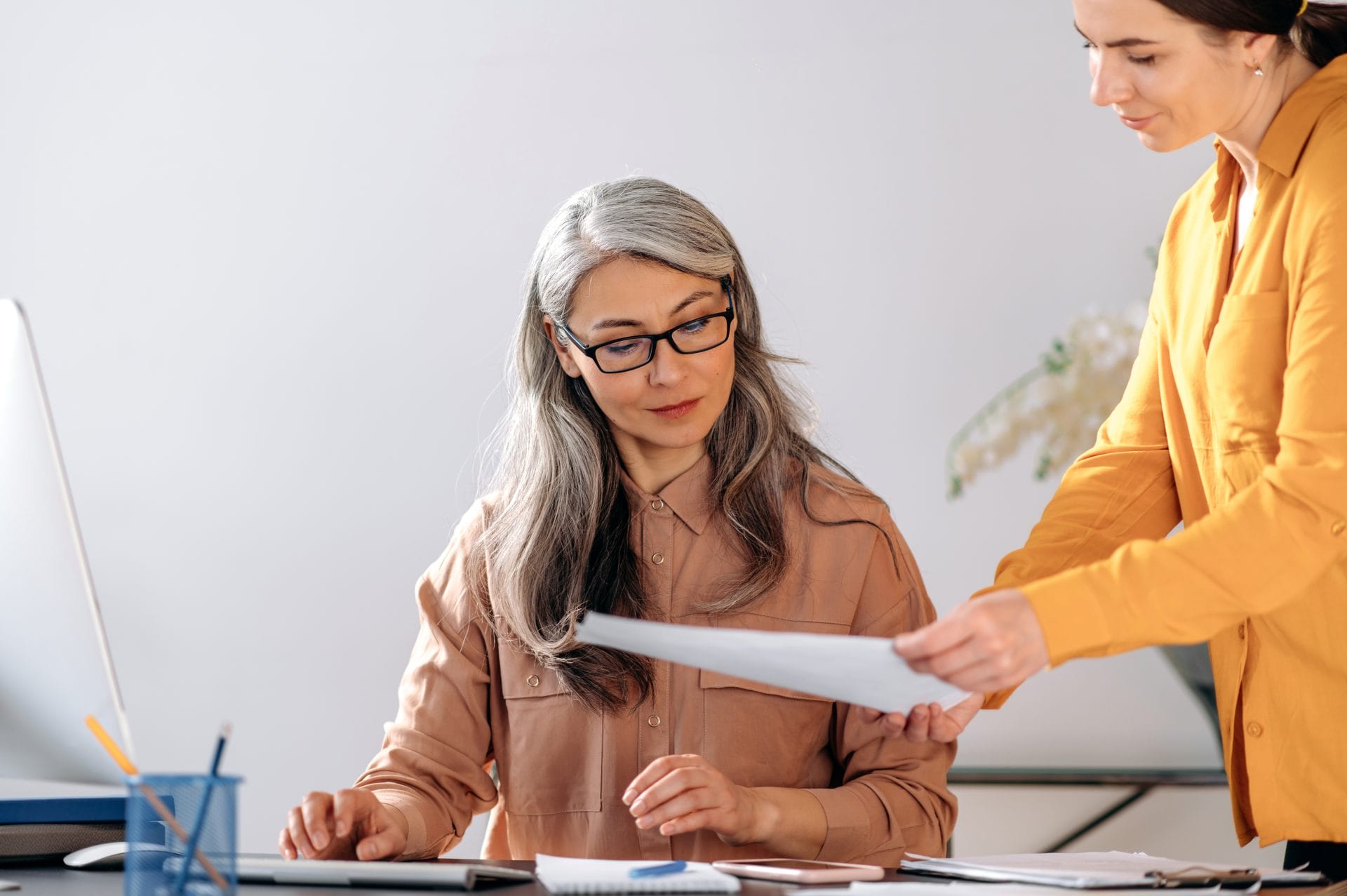 A gray-haired woman wearing glasses sits at a desk reviewing documents while another woman standing beside her hands her a sheet of paper. They appear to be working together in an office setting with a computer and notepads nearby.