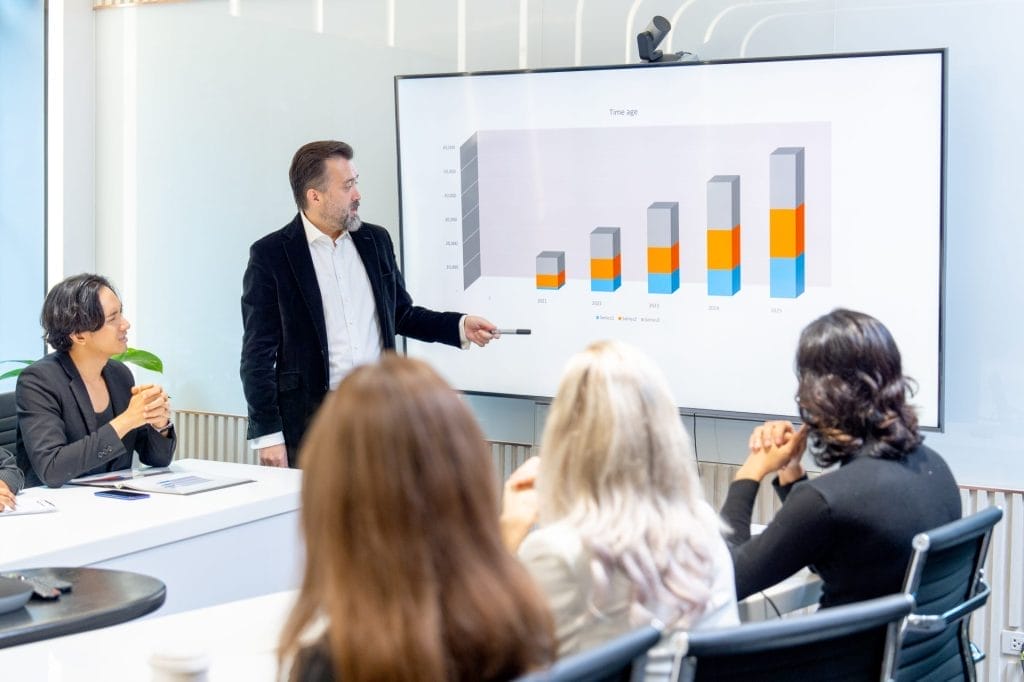 A presenter reviews performance data on a screen during a meeting, illustrating program outcomes and benchmarks.