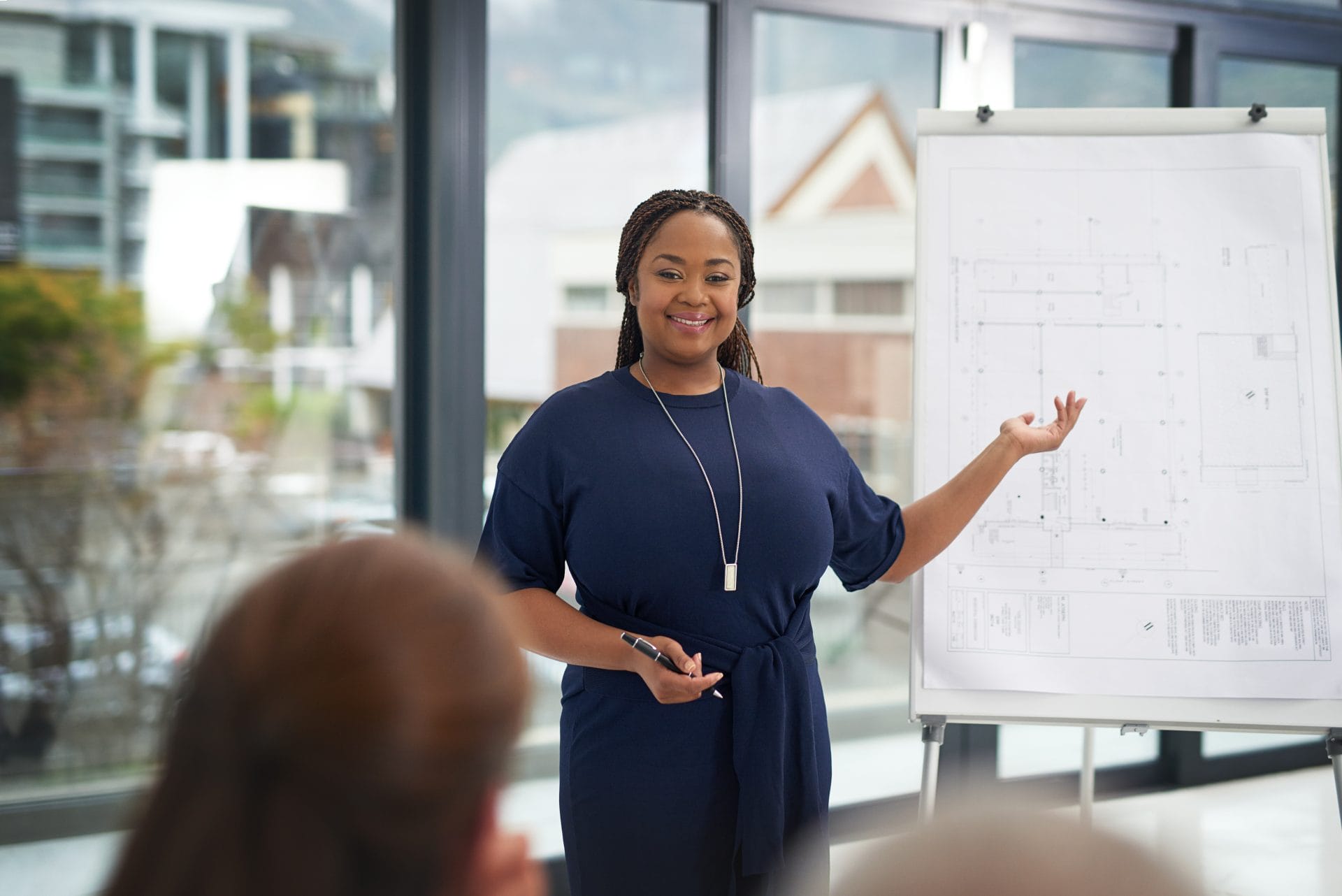A woman standing in front of a large flip chart, smiling as she gives a presentation to a small audience. She gestures toward architectural or technical drawings on the chart while holding a marker. Bright windows behind her show an urban landscape.