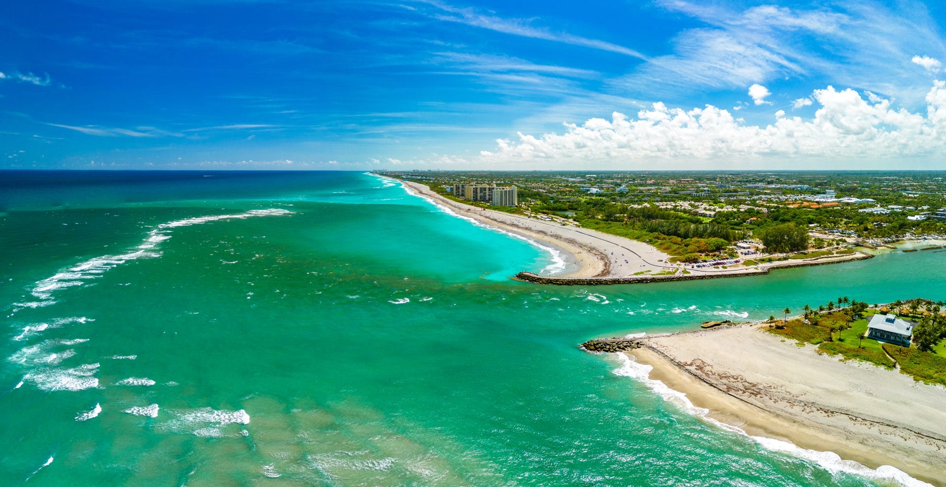 Aerial view of a coastal inlet and beach on a bright, sunny day. Turquoise water flows between two sandy shorelines, with gentle waves breaking along the coast. The shoreline curves into the distance, lined with greenery, a few buildings, and beach access areas. White clouds stretch across a vivid blue sky, highlighting the vibrant colors of the ocean and coastline.