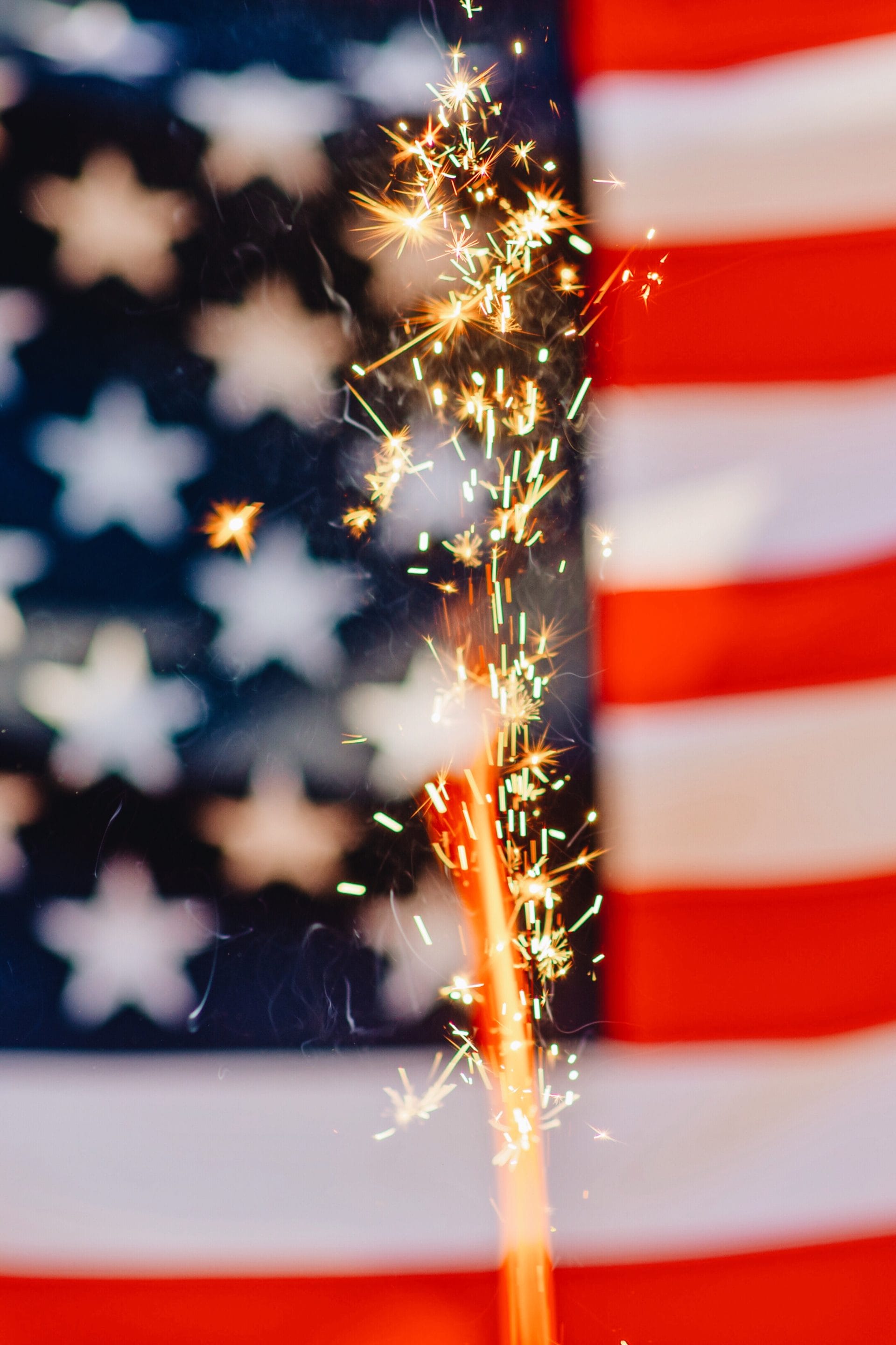 A close-up of a lit sparkler emitting bright golden sparks in front of a softly blurred American flag, creating a patriotic Fourth of July scene with a red, white, and blue backdrop.