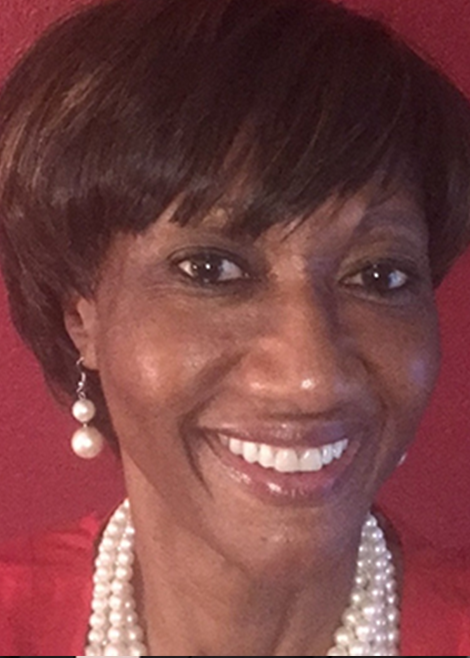 Professional headshot of a smiling woman with short dark hair wearing pearl earrings and a pearl necklace against a red background.