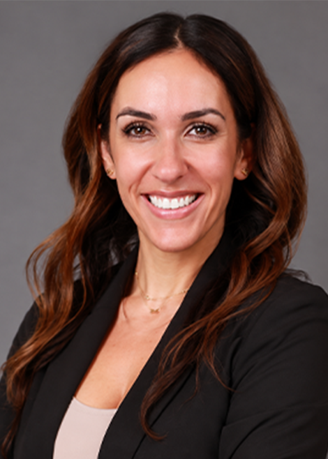 Professional headshot of a smiling woman with long wavy brown hair wearing a black blazer and beige top against a gray background.
