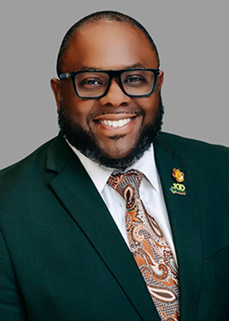 Professional headshot of a smiling man wearing black glasses, a dark green suit and a patterned tie against a gray background.